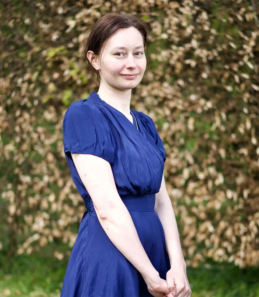 Photo of S.L. Dove Cooper in a blue dress against a background of green and brown vegetation.