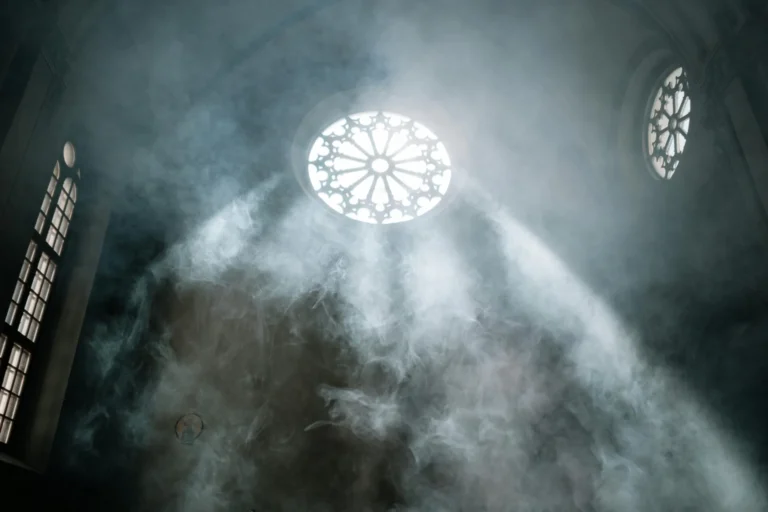 Photo of light shining through a round decorative window in a cavernous, ligtly smoke-filled space reminiscent of a church. Photo by cottonbro studio.
